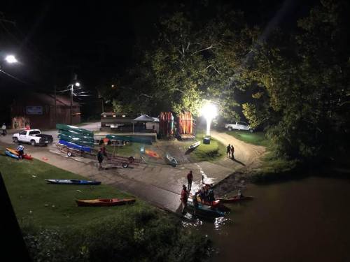 A nighttime scene at a boat launch with kayaks, canoes, and people preparing to launch into the water.