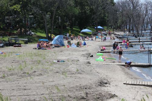 A busy beach scene with people sunbathing, playing, and relaxing near the water, surrounded by trees and tents.