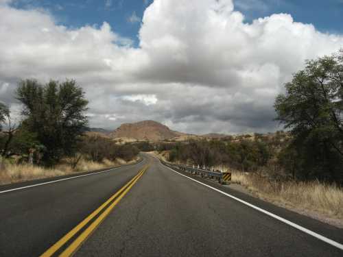 A winding road stretches through a dry landscape with hills and cloudy skies in the background.