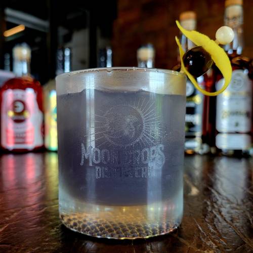 A cocktail in a clear glass with a lemon twist and a cherry garnish, set against a backdrop of liquor bottles.