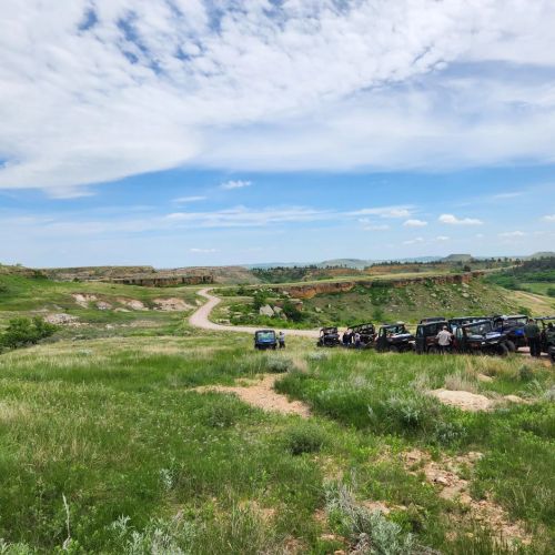 A scenic landscape with green hills, a winding dirt road, and parked off-road vehicles under a partly cloudy sky.