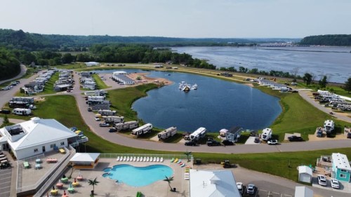 Aerial view of an RV park with a pond, pool, and river in the background, surrounded by green hills.
