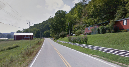 A rural road lined with greenery, leading past a red barn and a house, surrounded by hills under a partly cloudy sky.