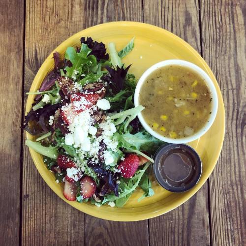 A yellow plate with a fresh salad topped with strawberries and feta, alongside a bowl of soup and a dressing cup.