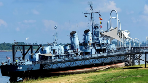 The USS Kidd DD 661, a blue naval destroyer, docked by the river with a clear sky in the background.
