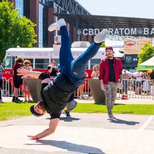 A person performs a breakdance move outdoors, with a food truck and spectators in the background.