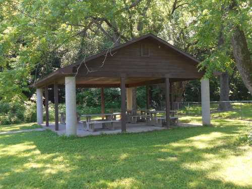 A wooden picnic shelter with a sloped roof, surrounded by trees and grass in a park setting.