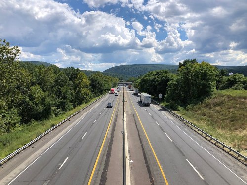 A view of a highway with vehicles traveling, surrounded by green trees and mountains under a cloudy sky.