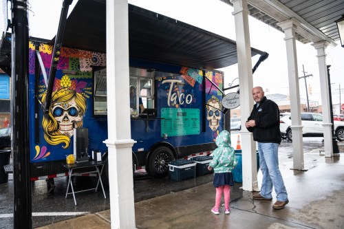 A colorful taco food truck under a covered area, with a man and a child standing nearby in the rain.