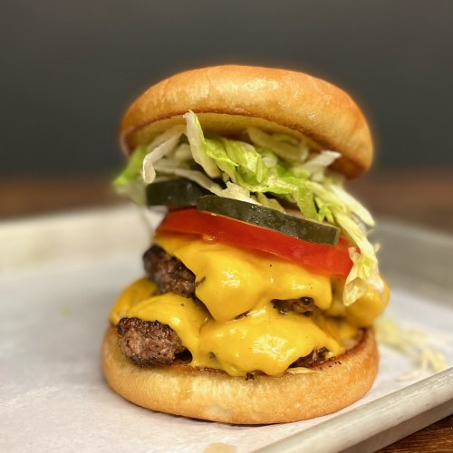 A stacked cheeseburger with two patties, melted cheese, lettuce, tomato, and pickles on a soft bun.