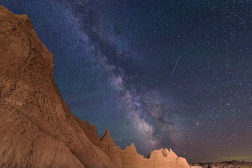 A starry night sky filled with the Milky Way over rugged, illuminated rock formations.