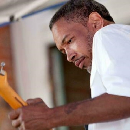 A man in a white shirt plays a guitar, focused on his performance outdoors.