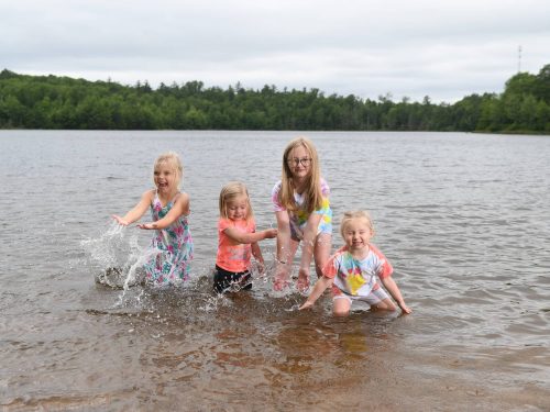 Four young girls play in shallow water, splashing and laughing, with trees and a cloudy sky in the background.