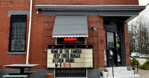 Sign outside a brick building advertising "Home of the Famous Deep Fried Burger" with kitchen hours listed.