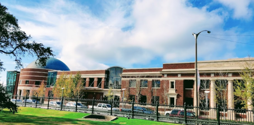 A modern brick building with a dome, surrounded by trees and a grassy area under a partly cloudy sky.