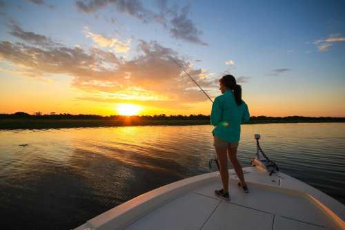 A person stands on a boat at sunset, fishing with a rod, surrounded by calm waters and a colorful sky.