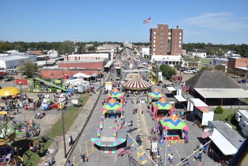 Aerial view of a vibrant street fair with colorful rides, booths, and a clear blue sky. American flags are visible.
