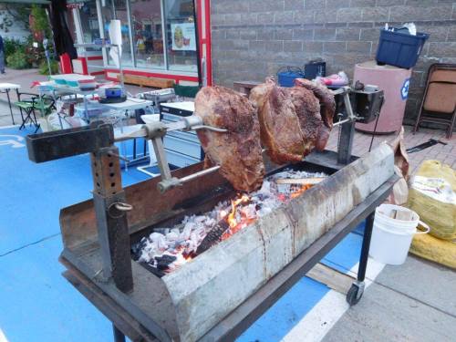 A large rotisserie grill with three pieces of meat cooking over an open flame, set up outdoors.