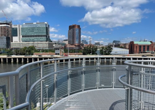 View of a city skyline with modern buildings and a river, featuring a circular metal walkway in the foreground.