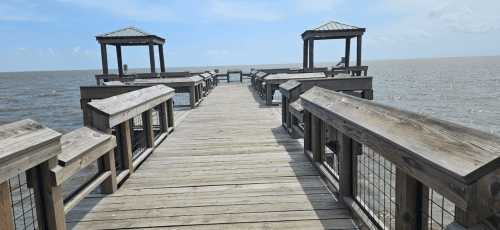 A wooden pier extends over calm water, featuring two covered viewing platforms on either side under a clear blue sky.