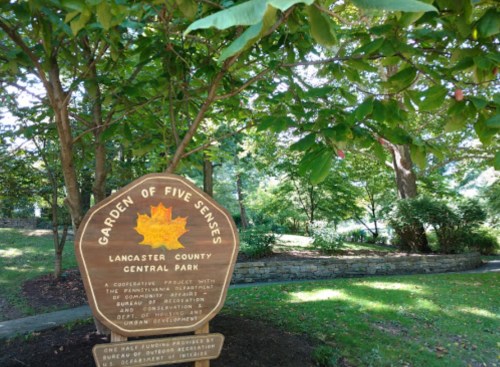 Sign for the "Garden of Five Senses" in Lancaster County Central Park, surrounded by lush greenery and trees.
