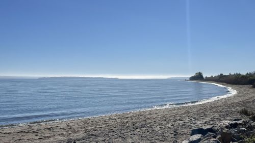 A serene beach scene with calm water, gentle waves, and a clear blue sky in the background.