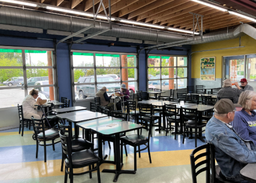 A spacious, brightly lit café with empty tables and a few patrons seated, enjoying their time by large windows.
