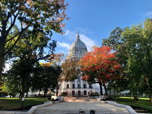 A view of a grand capitol building surrounded by colorful autumn trees and a clear blue sky.