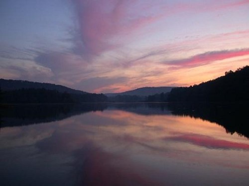 A serene lake at sunset, reflecting colorful clouds and surrounding hills in calm waters.