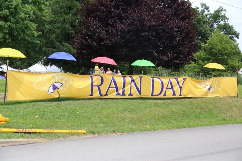 A large yellow banner reading "RAIN DAY" with colorful umbrellas in the background, set in a green park.