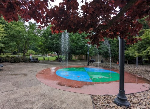 A colorful splash pad with water fountains, surrounded by green trees and benches in a park setting.