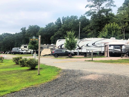 A gravel RV park with several parked trailers and a sign for "Linc Out Trail" surrounded by trees.