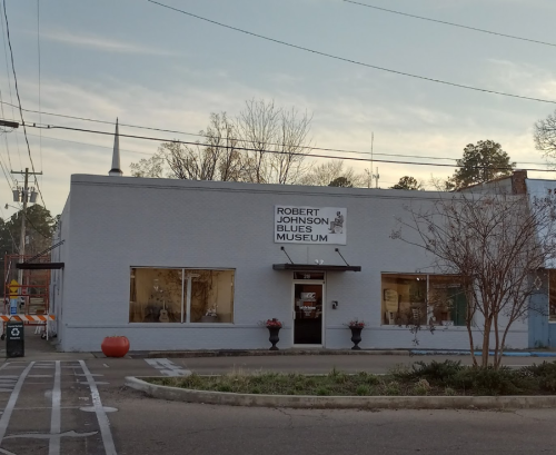 Exterior of the Robert Johnson Blues Museum, featuring a sign and large windows, with trees and a church steeple in the background.
