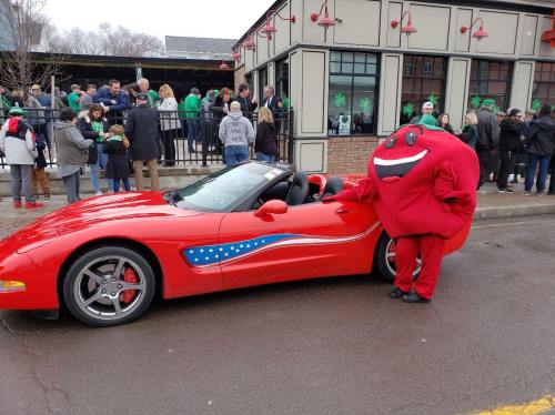 A red sports car with a patriotic design parked on the street, next to a person in a large red mascot costume.