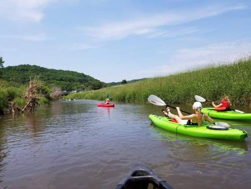 Three people kayak on a calm river surrounded by lush greenery and hills under a clear blue sky.