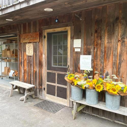 A rustic bakery entrance with a wooden door, surrounded by flower pots filled with sunflowers and greenery.