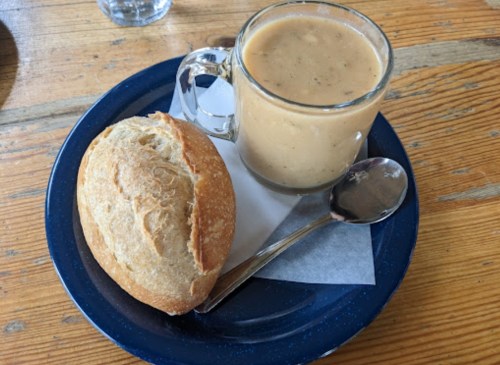 A glass mug of soup beside a crusty roll on a blue plate, with a spoon and napkin on a wooden table.