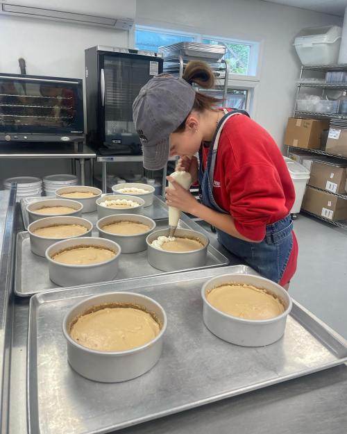 A person in a red sweater and cap pipes frosting onto cheesecake in a kitchen with multiple cake pans.