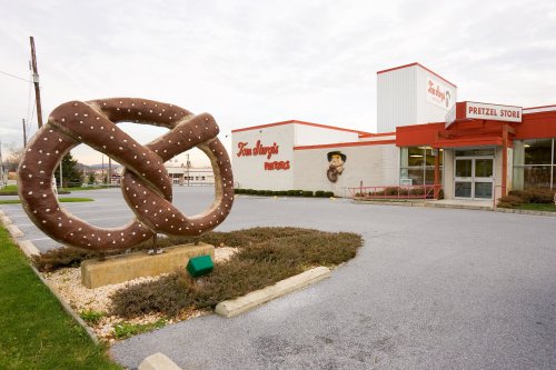 A large pretzel sculpture stands outside a pretzel store with a mural on the building and a cloudy sky above.