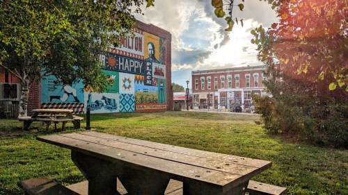 A park scene featuring a wooden picnic table, vibrant mural on a building, and a sunny sky with scattered clouds.