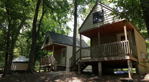 Two wooden cabins nestled among trees, featuring porches and a yurt nearby, in a serene outdoor setting.