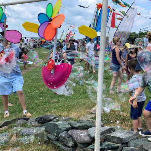 A lively outdoor scene with people enjoying a festival, colorful kites flying, and bubbles floating in the air.