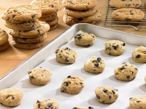 A tray of cookie dough balls on parchment paper, with baked cookies stacked in the background.