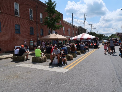 A crowd gathers on a street with tents and hay bales, enjoying a sunny day at a community event.