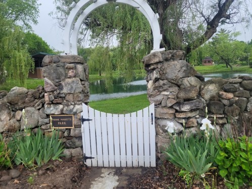 A white wooden gate in a stone archway, leading to a lush green park with a pond and trees in the background.