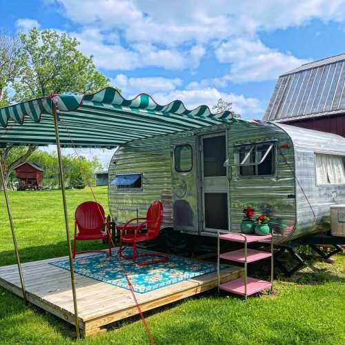 A vintage silver trailer with red chairs and a green striped awning, set on a wooden deck in a grassy area.