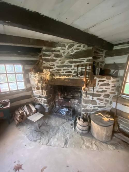 A rustic stone fireplace in a log cabin, surrounded by wooden beams and various kitchen tools.