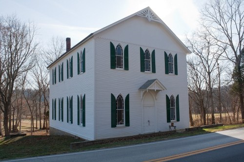 A white, historic building with green shutters and pointed windows, set against a backdrop of bare trees and a clear sky.