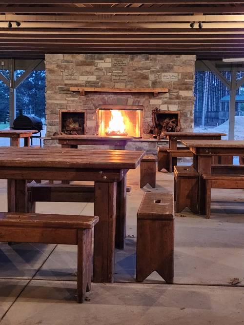 Cozy outdoor seating area with wooden tables and a stone fireplace glowing with fire.