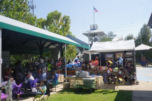 A sunny outdoor market scene with tents, vendors, and people browsing various goods. An American flag flies above.